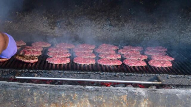 Dozens of hamburger patties sizzle and smoke over glowing hot coals on a large, open outdoor barbecue grill as a cook wearing a purple nitrile glove flips them.
