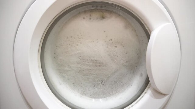 the washing machine washes white laundry with plenty of foam inside the machine. view through the round glass of the washing machine.