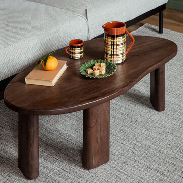 Cozy modern living room interior with a dark wood coffee table, ceramic jug, mug, book, and fresh orange on a soft grey rug