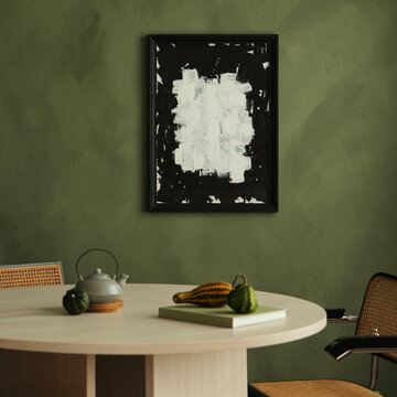 Minimalist dining room interior with a round table, green wall, abstract art piece in a black frame, ceramic teapot, and small gourds