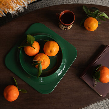 Fresh mandarins with green leaves in a dark green plate on a wooden table alongside a cup of coffee and a book top view flat lay scene
