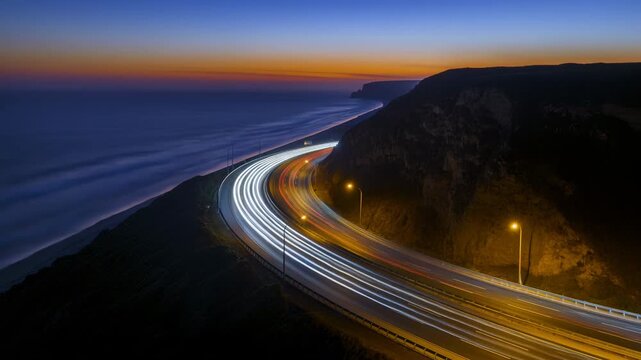 Coastal highway curves along cliffside coastline as vehicle light trails stream through winding road at dusk with ocean view and dark landscape creating dynamic travel motion scene