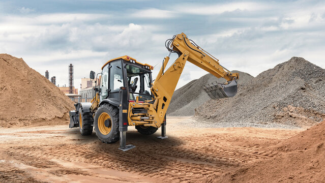Excavator loader is digging and moving dirt at a construction site with piles of gravel and sand in the background on a cloudy day in the city