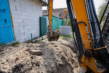 A digger is lifting dirt at a construction site next to a building. Workers are preparing the area for future work. The sun is shining overhead © Anoo
