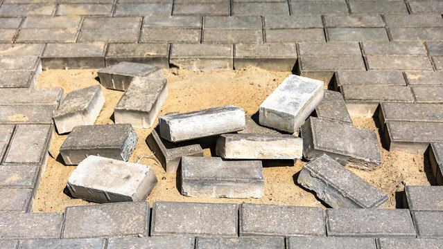Bricks lay scattered on sand in a gap of a paved surface. Nearby, the surrounding bricks align neatly. This scene takes place during a construction project