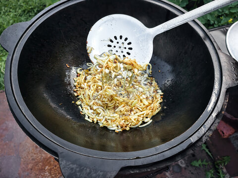 In a garden, a person stirs fried onions and spices in a kazan over an open flame, preparing plov for a gathering