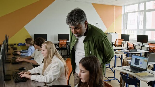 Middle aged South Asian teacher guiding diverse teenagers working on computers in school lab. Ideal for promoting STEM education, digital literacy, and modern school curriculum advertisements