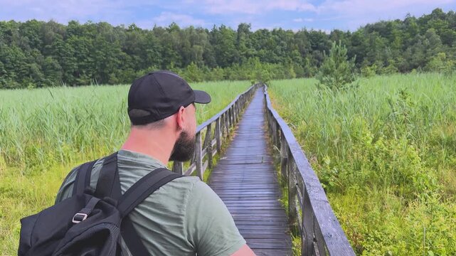 Bearded male hiker walking on a wooden boardwalk through a swamp and turning back to smile at the camera