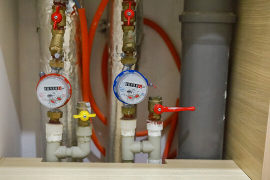 Hot and cold water meters with pipes, valves and hoses are installed in a residential plumbing cabinet. Utility metering represents water consumption monitoring and plumbing maintenance.