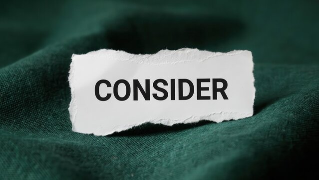A torn white paper strip with the word consider printed on it rests on a dark green textured fabric. Ideal for decision making, thinking, and business strategy concepts.