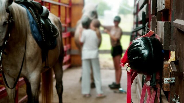 Riding helmet and horse tack hang inside stable beside saddled gray horse. Children care for equine in blurred background.