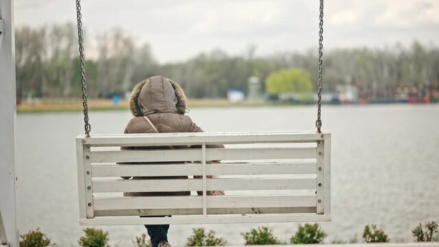 Person sitting alone on wooden swing bench facing lake view in park.