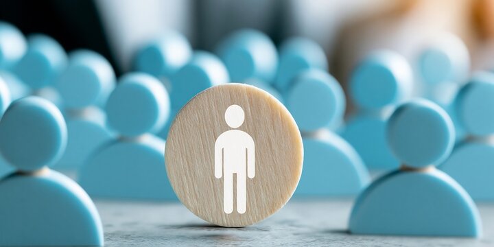 Wooden token with white human icon in sharp focus, set before a blurred crowd of blue person-shaped figures; concept of individuality, leadership, and human resources