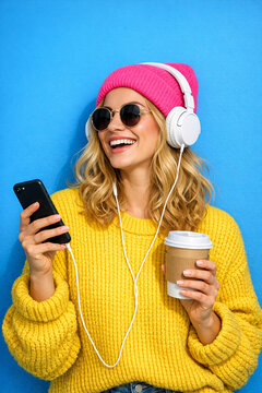 A modern woman with blonde hair, wearing a yellow sweater and pink beanie, listening to music while holding her smartphone and coffee cup.