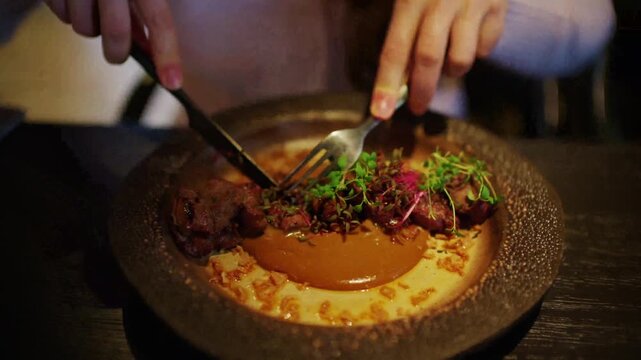Person using silverware to cut and eat gourmet meat dish at upscale restaurant.