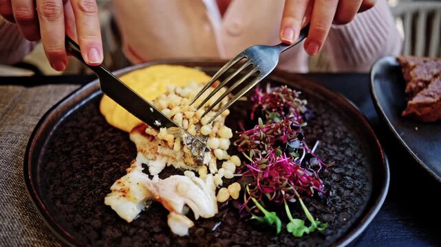 Woman using knife and fork to eat gourmet white fish dish with corn and microgreens.