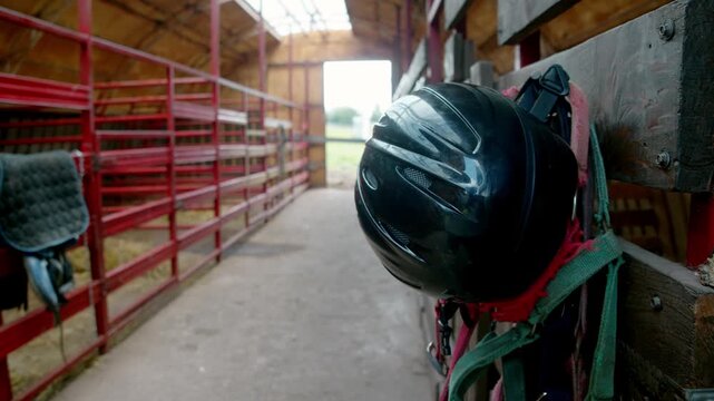 Black riding helmet hangs with colorful horse tack inside a stable aisle. Red stall rails and saddle equipment create an equestrian setting.