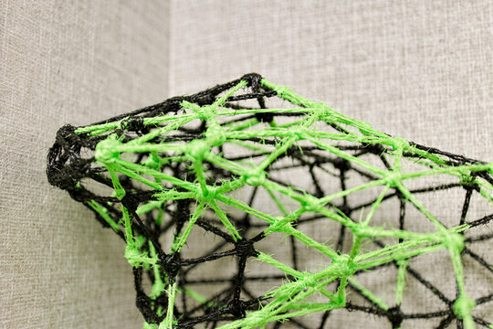 A modern geometric 3D wireframe mesh sculpture created with neon green and black knotted string cords against a neutral textured wall