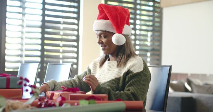 Mature woman in Santa hat preparing holiday gifts, wrapping boxes, tying ribbons at dining table