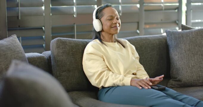 African American woman closing eyes, leaning back on sofa, touching remote, relaxing with headset