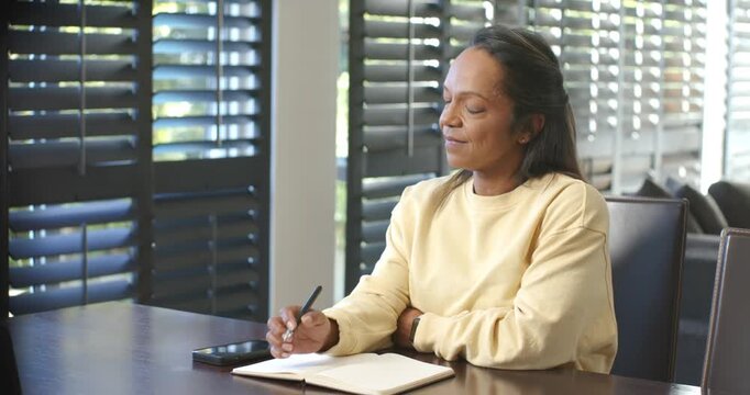 Mature African-American woman writing with pen in journal at table by shutters thinking, copy space