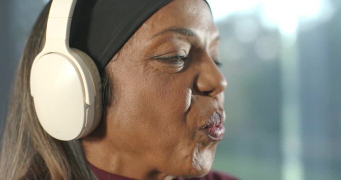 Senior African American woman hearing audio on white headphones, headband singing smiling by window