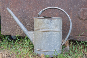Vintage galvanized watering can placed on green grass beside a weathered rusty metal garden structure © Olha