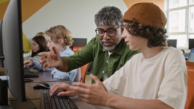 Middle-aged South Asian teacher explaining computer programming to teenage student typing on keyboard in classroom. Footage is ideal for promoting STEM education and coding bootcamps