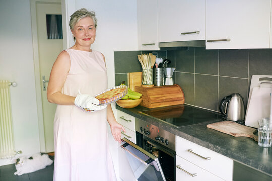 55-60 Year Old Woman Holding Fresh Homemade Pie by Open Oven in Modern Kitchen, Smiling Mature Female Enjoying Baking and Cooking at Home