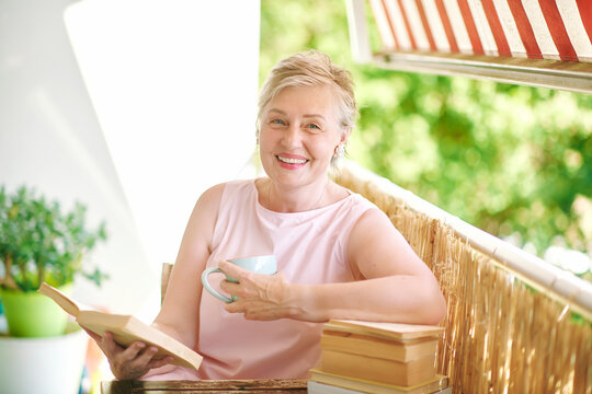 woman in her 50s reading a book and holding a cup while relaxing on sunny balcony at home, smiling mature female enjoying quiet leisure time and reading