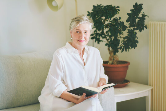 55-60 year old woman reading a book while sitting on sofa at home, relaxed mature female enjoying quiet leisure time, literature and peaceful lifestyle