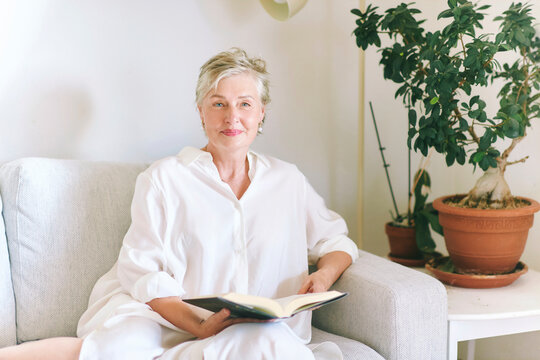 55-60 year old woman sitting on sofa with an open book at home, smiling mature female enjoying reading, quiet leisure time, comfort and peaceful lifestyle