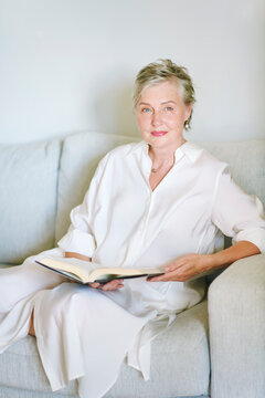 55-60 year old woman sitting on sofa with an open book at home, elegant mature female relaxing and enjoying reading, peaceful leisure time and quiet lifestyle