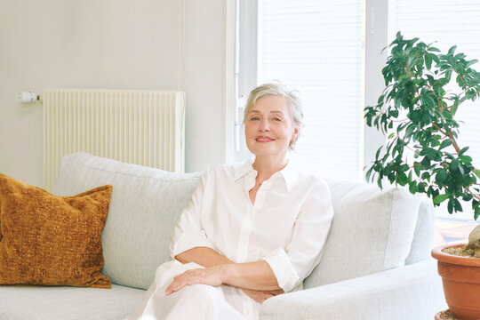 55-60 year old woman sitting comfortably on sofa at home, smiling mature female in white clothes relaxing in bright living room, confident and peaceful lifestyle