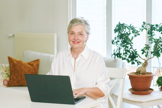 55-60 year old woman using laptop computer while sitting at home, smiling mature female working online, browsing internet and enjoying modern digital lifestyle