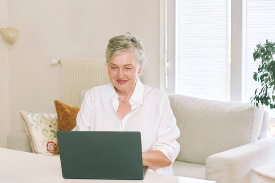 55-60 year old woman using laptop computer while sitting at home, smiling mature female working online, browsing internet and enjoying modern digital lifestyle