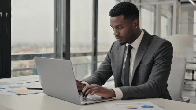 A man dressed in a suit and tie works on his laptop in a modern office with a view.