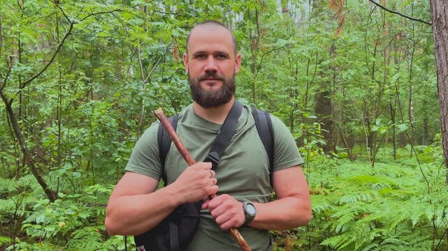 Portrait of a bearded male hiker with backpack and walking stick standing in a green forest looking at camera