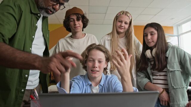 Multiethnic middle aged teacher guiding Caucasian teenagers collaborating on laptop in classroom. Ideal for marketing STEM education, coding bootcamps, and modern school learning programs