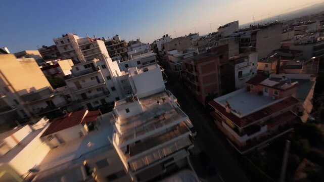 Dynamic fast-paced FPV drone footage diving past residential rooftops and apartment buildings in an urban Athens neighborhood facing Lycabettus Hill and the warm golden flare of sunset.