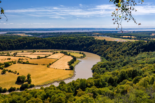 River Wye winding past dry golden fields and dense woodland near Chepstow, Severn Estuary and suspension bridge visible across distant horizon, Monmouthshire, Wales
