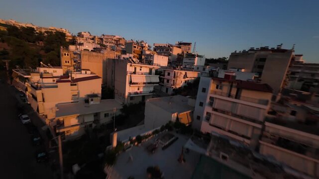 Dynamic fast-paced FPV drone footage diving past residential rooftops and apartment buildings in an urban Athens neighborhood facing Lycabettus Hill and the warm golden flare of sunset.