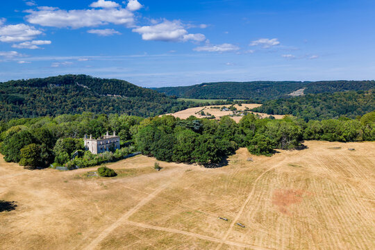 Abandoned 18th century country house ruins nestled among mature trees, yellow parched fields in foreground and wooded ridges under blue sky, Chepstow, Wales