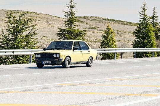 A yellow 1980s Tofaş Murat 131 classic car drives along a multi-lane highway against a backdrop of rolling hills and coniferous trees in Anatolia a classic cult car on the road in Konya, Turkey 2.4.25