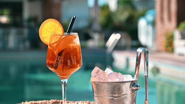 Aperol spritz cocktail glass with orange slice next to ice bucket by swimming pool in sunny day.