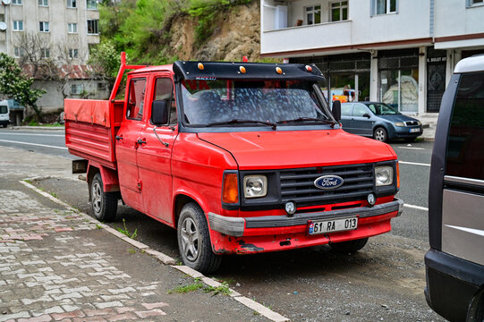 A red Ford Transit Mk2 double-cab flatbed truck with a sun visor is parked on the side of the road in Anatolia; a classic 1980s double-cab vintage truck on the road in Turkey