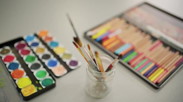 Creative art supplies including watercolor paint set and colorful drawing pencils arranged on tabletop for artistic hobby projects.