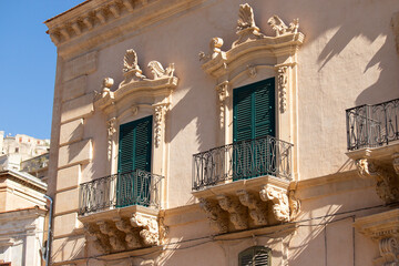 Modica, Italy  1st August 2026: Baroque architectural detail with carved grotesque face and ornate stonework on a balcony in Sicily. © Leckerstudio