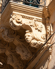 Modica, Italy  1st August 2026: Baroque architectural detail with carved grotesque face and ornate stonework on a balcony in Sicily. © Leckerstudio