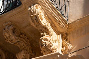 Modica, Italy  1st August 2026: Baroque architectural detail with carved grotesque face and ornate stonework on a balcony in Sicily. © Leckerstudio